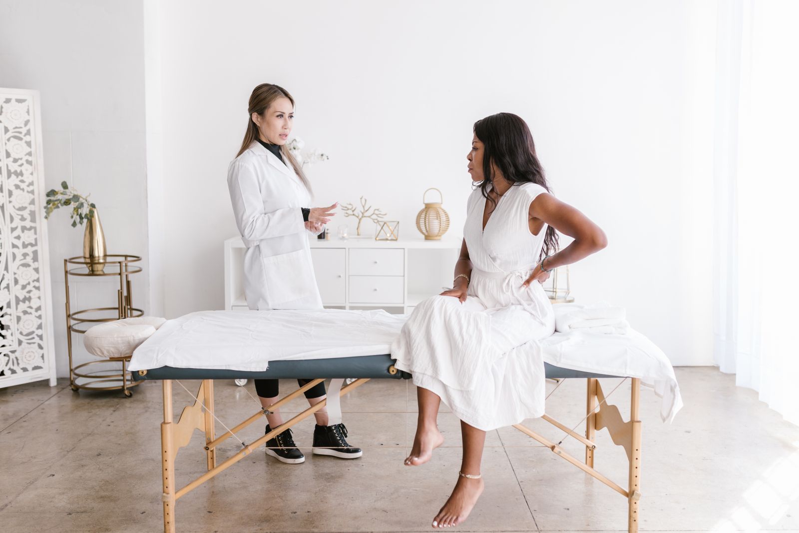 A patient lying face-down on a treatment table with a hand resting on her lower back, consulting with a clinician in a white coat.